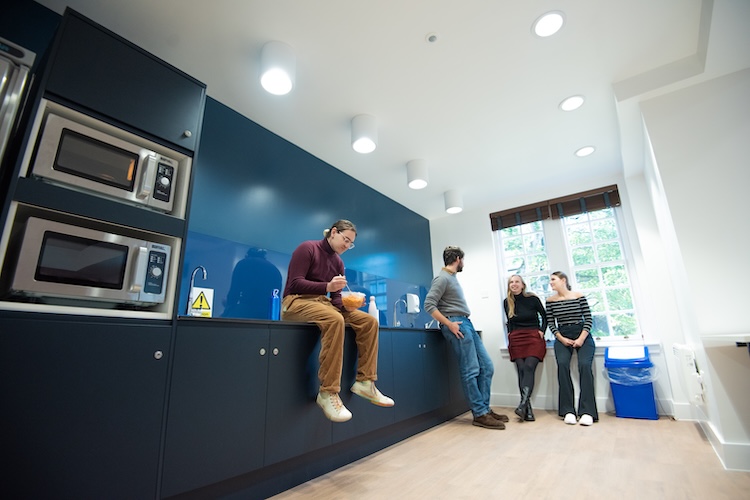 Four students chat in a shared kitchen, with one sitting on the counter holding a bowl.