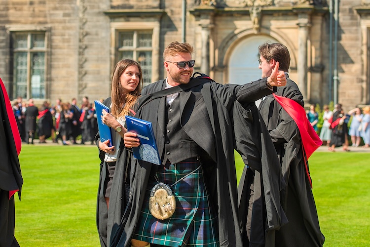 Graduate in academic robes and a tartan kilt gives a thumbs-up while standing with fellow graduates outside a university building.