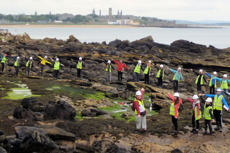 Students wearing hard hats and high-visibility vests explore a rocky shoreline, with the St Andrews skyline visible across the water.