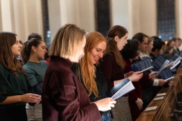 Two young women are holding paper programmes and standing in a church pew at a carol service. One is smiling.