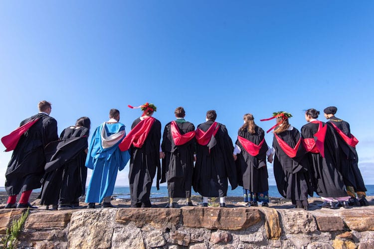 Ten graduates in gowns and hoods standing on St Andrews pier looking out to sea.