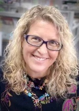 Headshot of Virginia Parker - a white woman with curly blonde hair and glasses, wearing a black top with a colourful necklace.