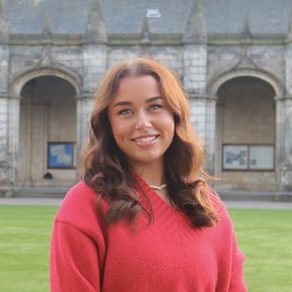 Missy facing 3/4 to camera wearing a red/pink jumper. She has long brown hair and is smiling. In the background is St Sally's quad.