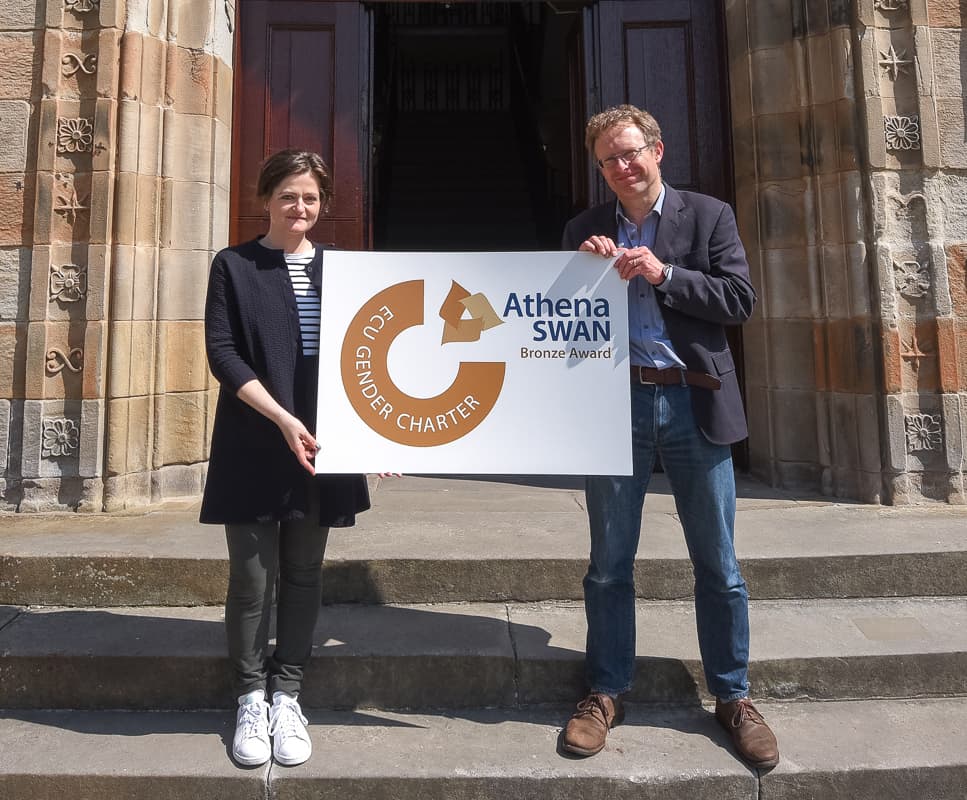 Two people holding a board with the Athena Swan logo on it