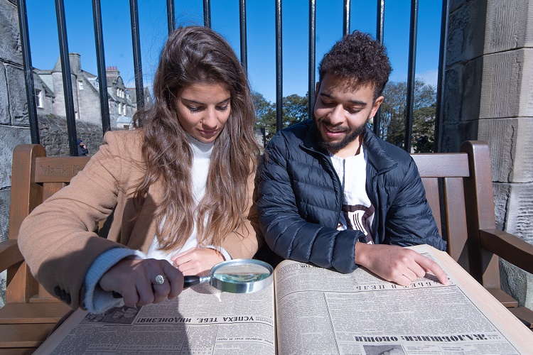 Two students examining a giant book with a magnifying glass