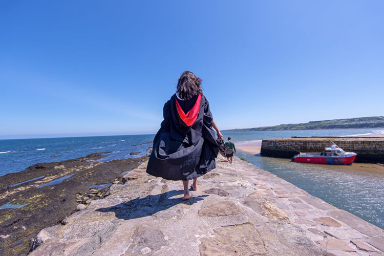 A femail student in a gown walking barefoot at St Andrews harbour