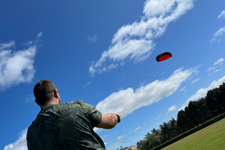 Mike flying a red and black kite in a blue sky with some fluffy white clouds.