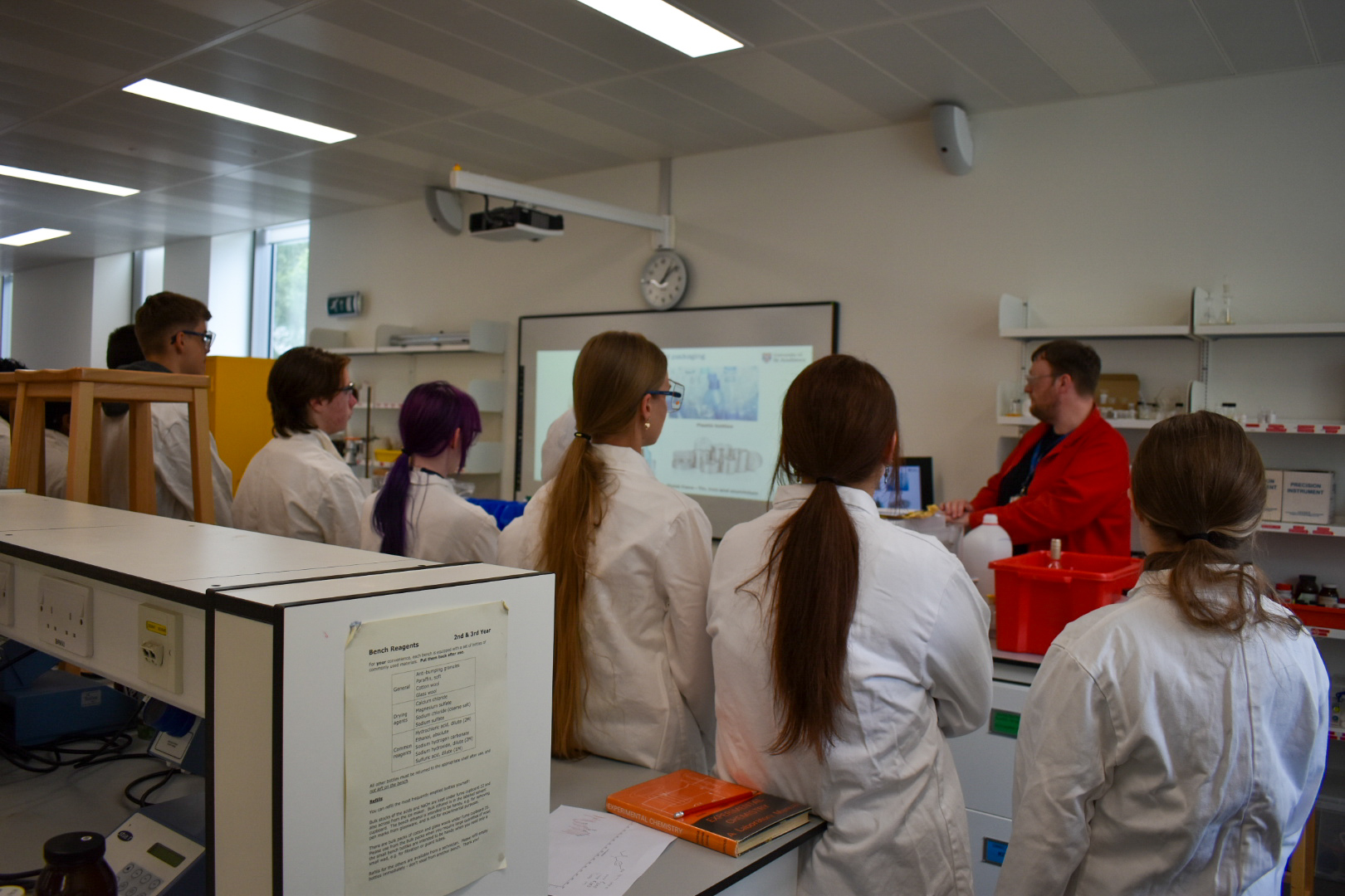 Five students in lab coats watching a lecture in a lab.