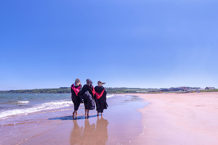 Students in graduation gowns walking along the beach