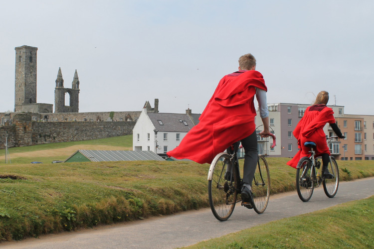 Two students in red gown are cycling along the path of East Sands Beach