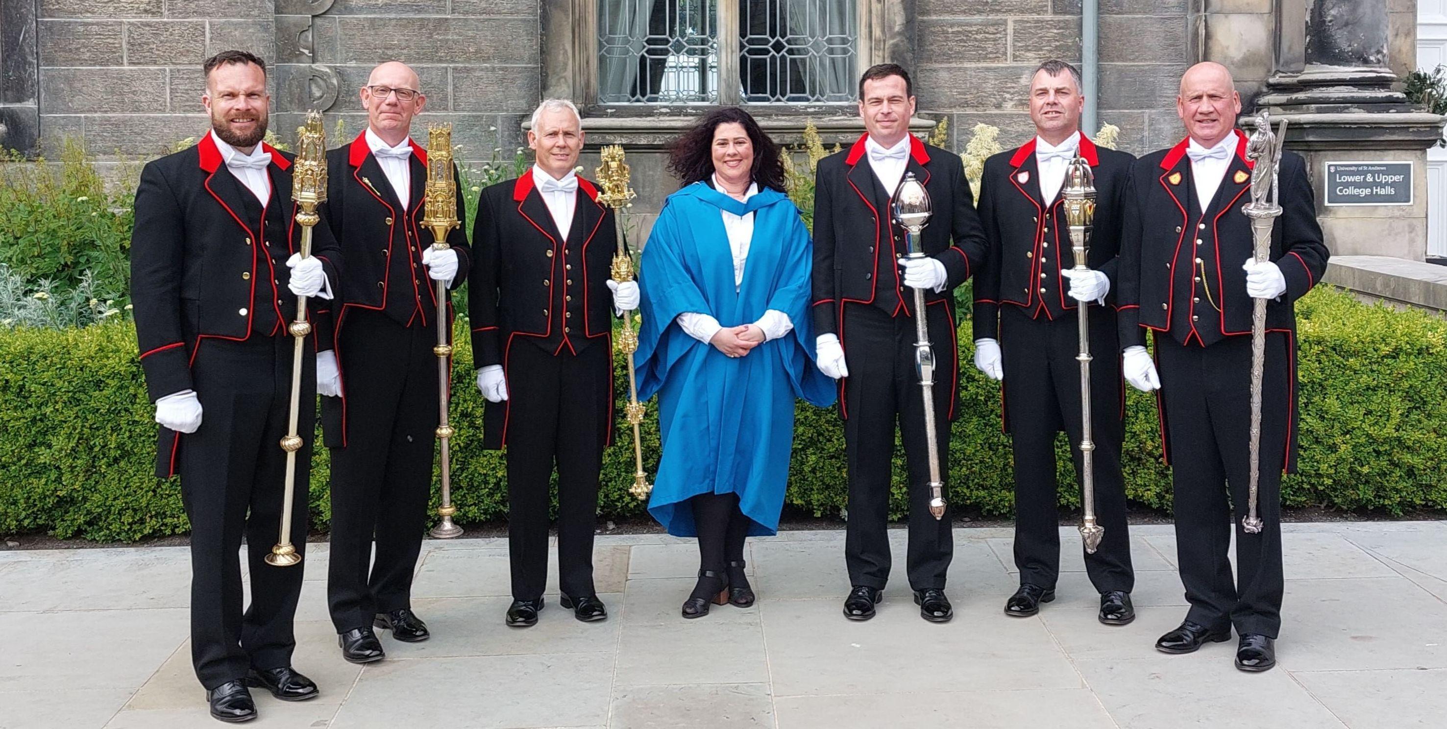 Six mace bearers in their formal attire pose outside Lower & Upper College Halls. Flavia smiles in the centre in a blue graduation gown.
