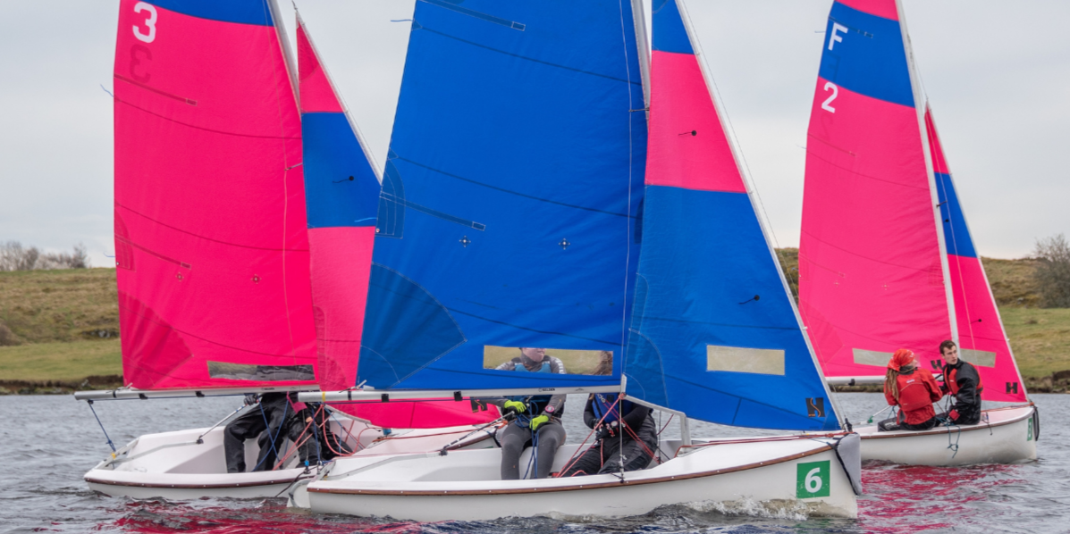 Three St Andrews Sailing Club dinghies can be seen in the water, each boat has two athletes aboard, the pink and blue sails are raised