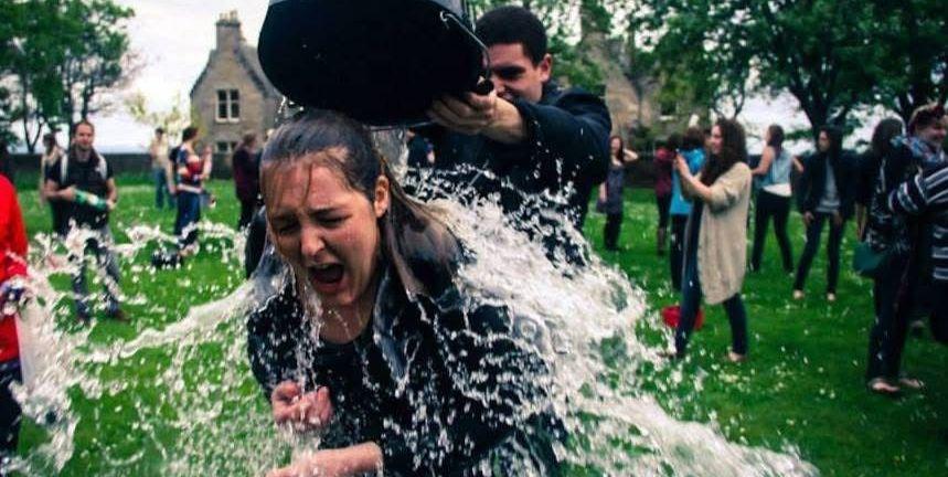 Collette being drenched with water from a black bucket, surrounded by people on a grassy area with trees. This is part of the traditional 'soaking' at St Andrews.