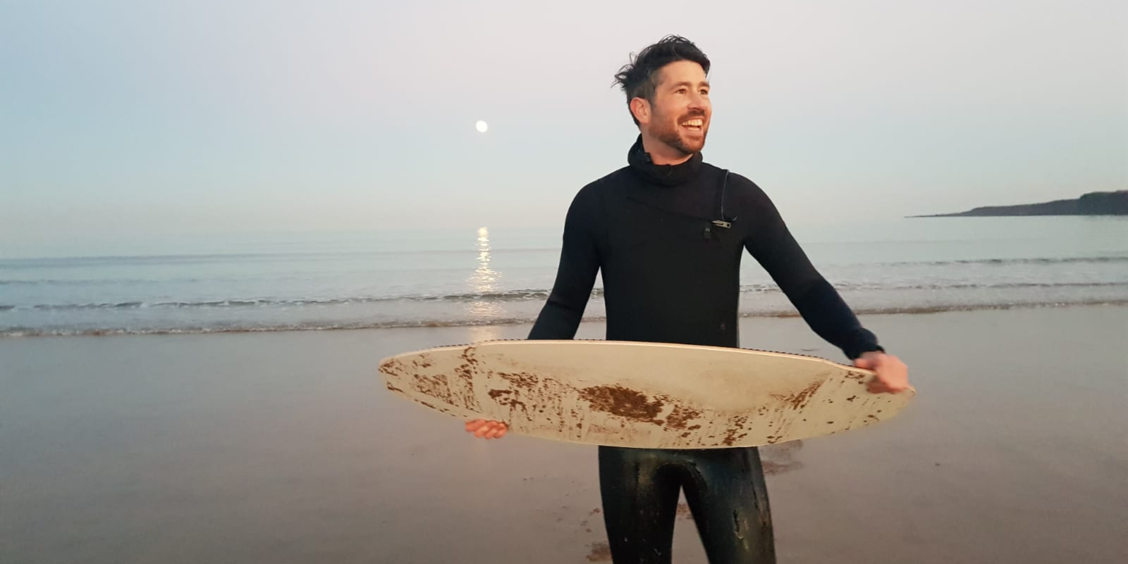 Dr Chris McKnight wears a black wetsuit holding a white surfboard on a sandy beach at sunset, with calm waves, the moon reflecting on the water. 