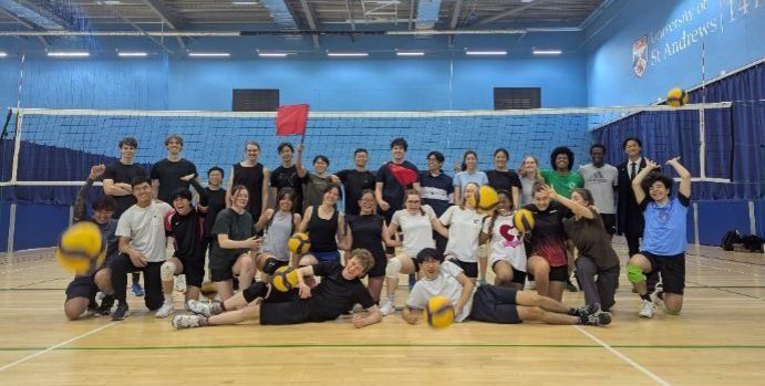 The University of St Andrews Volleyball Club pose for a team photo on the court. Volleyballs can be seen to be flying towards the camera. 