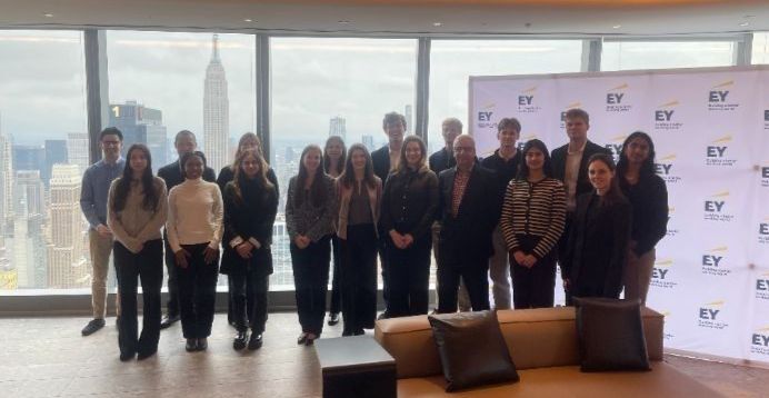 Group of students stand smiling with Jessica in front of EY branding and New York City skyline 
