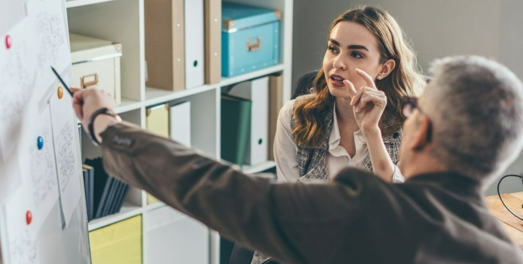 A person is pointing at a whiteboard covered with papers and colorful magnets while another person sits nearby, appearing to listen. The setting is an office space with shelves in the background holding boxes, binders, and storage containers.