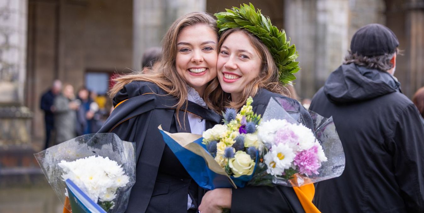 Two smiling graduates in their gowns pose for a photo in St Salvator's Quad, the graduates hold bouquets of flowers