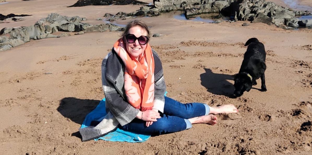 Dr Amy Blakeway sits on a beach towel on the sand, she smiles at the camera. Beside Amy there is a black Labrador. 