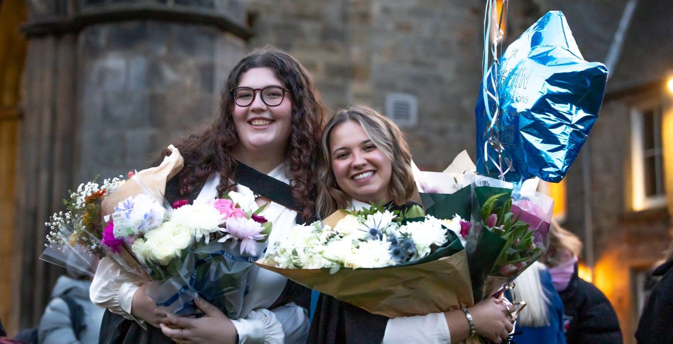 Two smiling graduates  pose for a photo on graduation day, the graduates hold bouquets of flowers - one holds a blue star balloon 