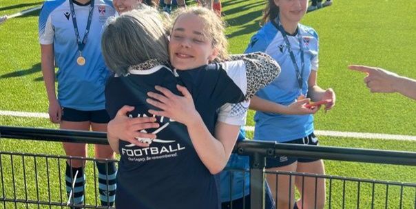 A grandmother, Fiona Goodchild, hugs her granddaughter as they celebrate a win for her football team. The young female footballers are in a blue strip on the pitch. A grandmother, Fiona Goodchild, hugs her granddaughter as they celebrate a win for her football team. The young female footballers are in a blue strip on the pitch.
