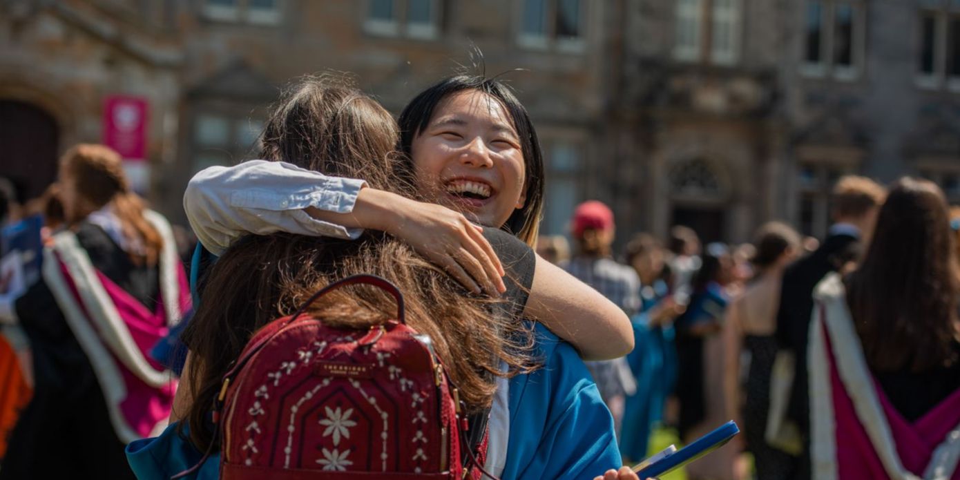 Two women embrace in a hug, one of the pair a graduate in blue robes and is smiling facing the camera. In the background a busy St Salvator's Quad can be seen in the background