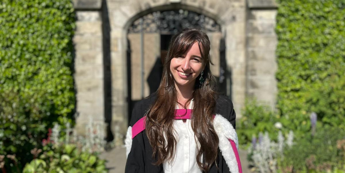 Patricia stands smiling in her graduation gown and academic dress, she is in St Salvator's Quad on a sunny day