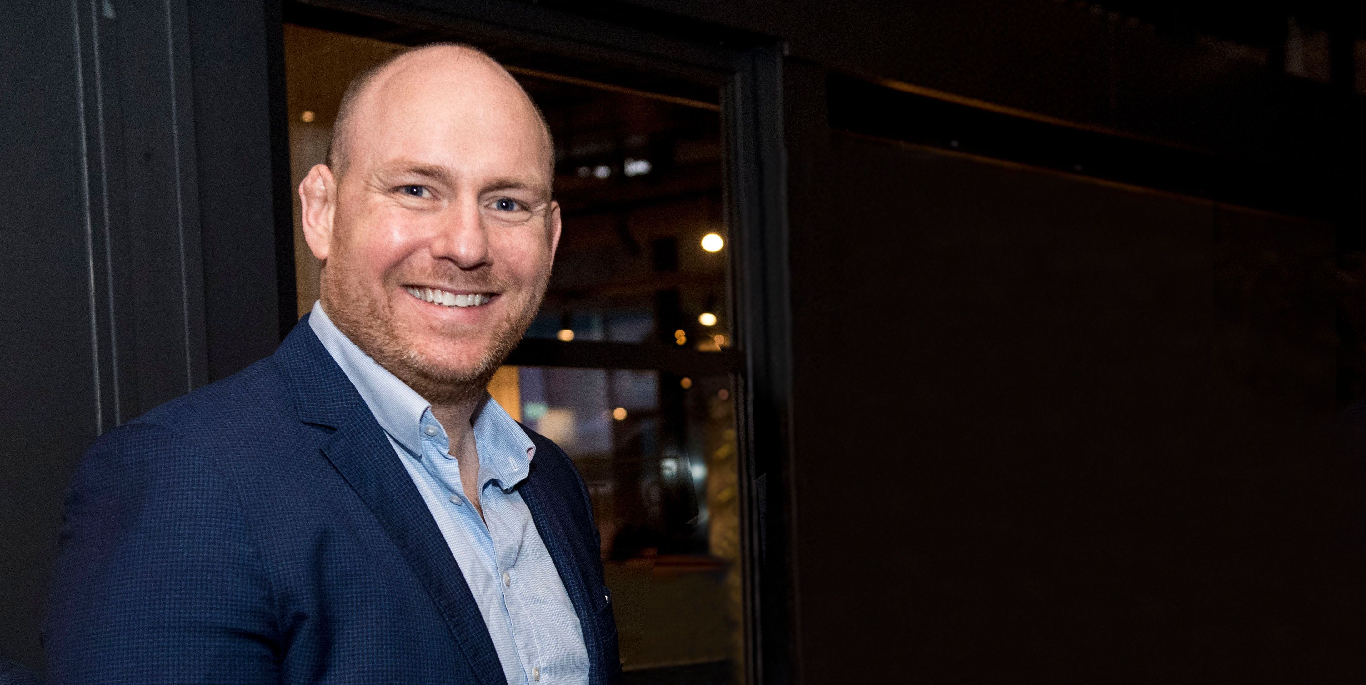 Peter stands smiling for a photo, he is dressed in a navy blue suit with a blue shirt