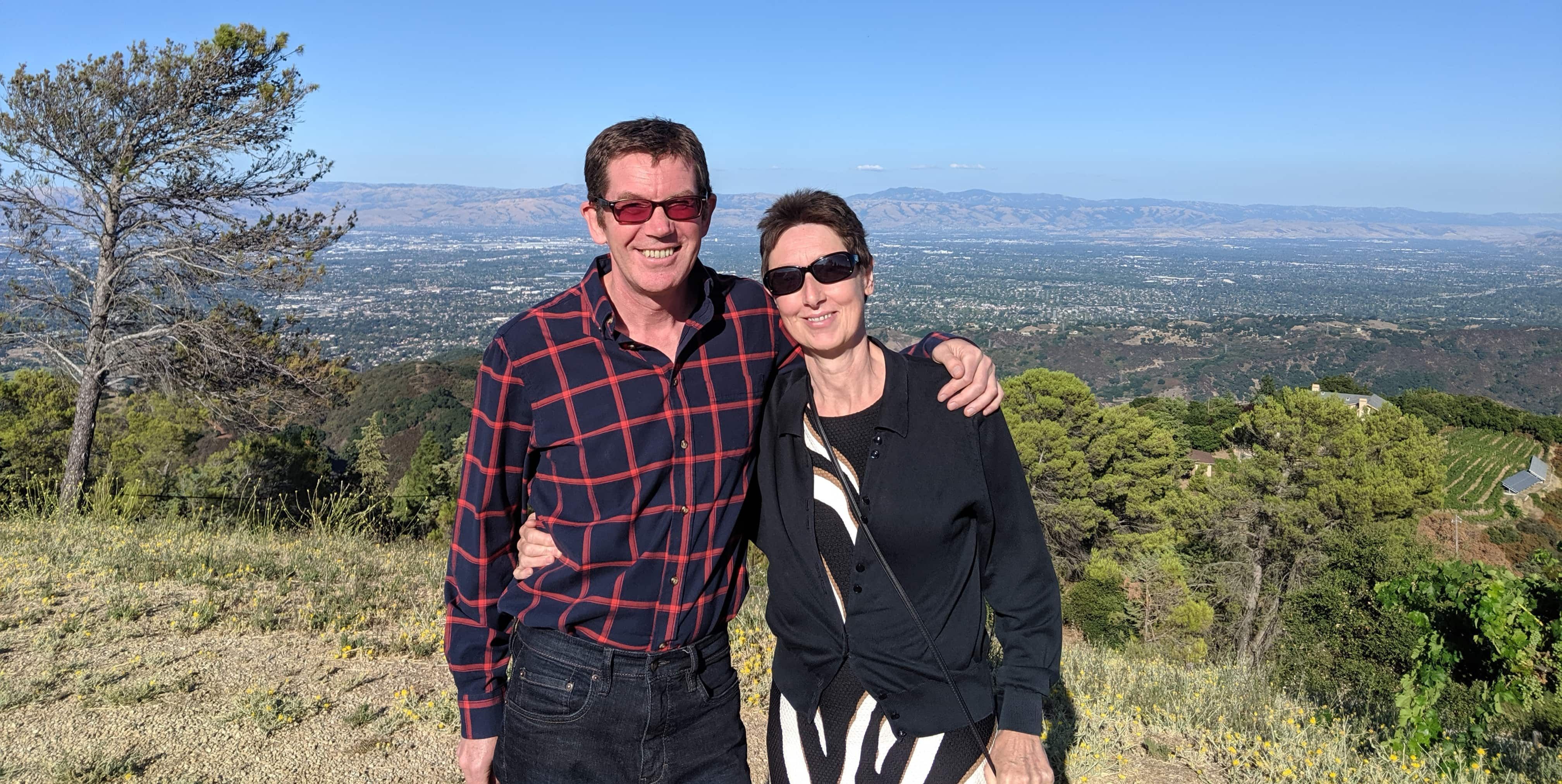 A couple, Lindsay and Nicky Grant, stand atop a hill in Silicon Valley, blue skies and vineyards behind them.