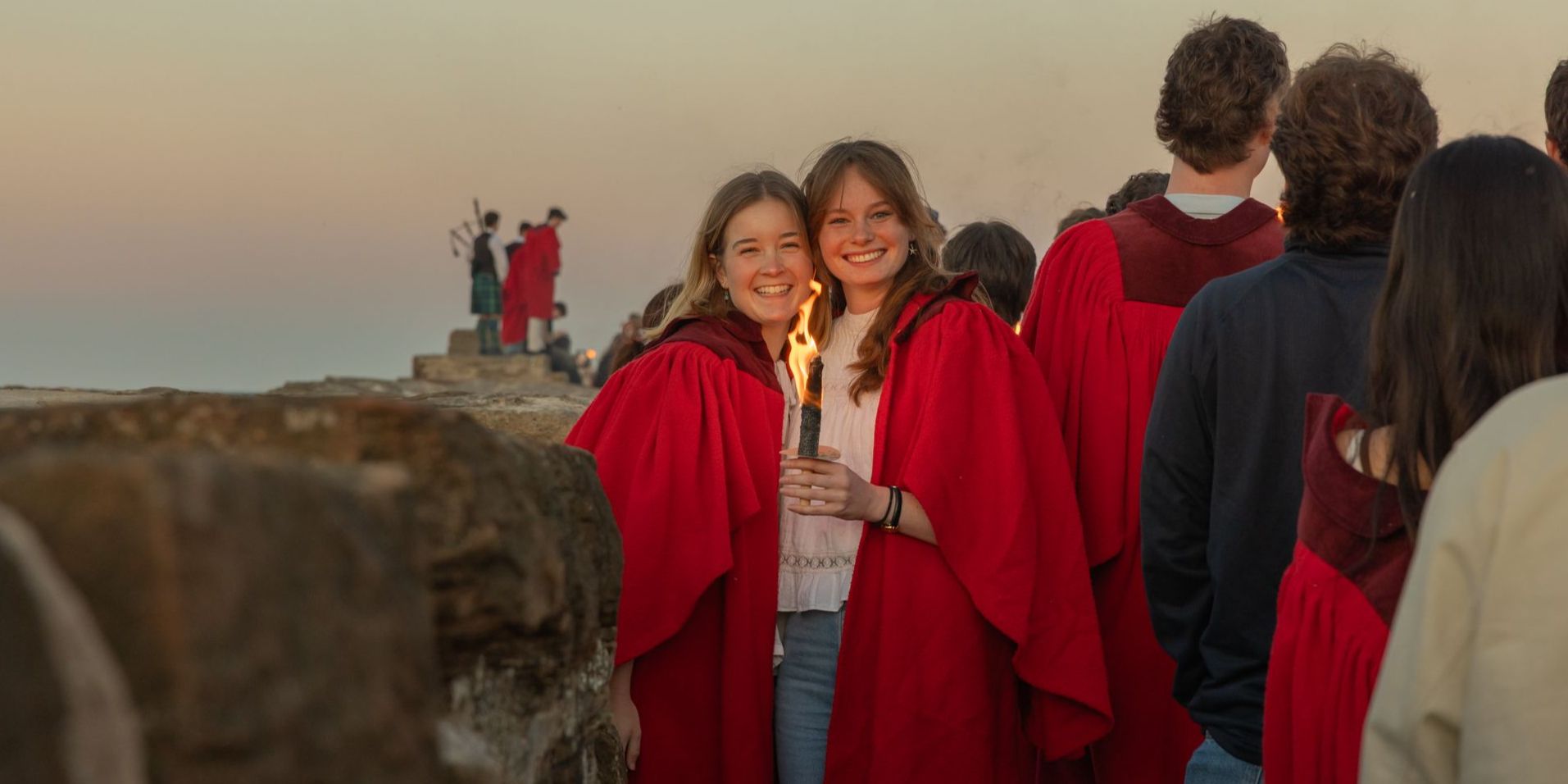 Two students stand on the St Andrews pier during the gaudie, both are wearing red gowns and one hold a torch. To the right of the pair, a crowd of fellow students participating in the Gaudie. To the left, a piper and two students in red gowns stand on the end of the pier