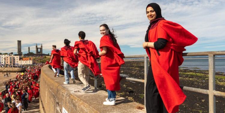Two windswept students look back smiling at the camera from the top of St Andrews Pier. They are two of many students participating in the Gaudie, wearing red gowns. A large group of participants can be seen on the pier below Two windswept students look back smiling at the camera from the top of St Andrews Pier. They are two of many students participating in the Gaudie, wearing red gowns. A large group of participants can be seen on the pier below