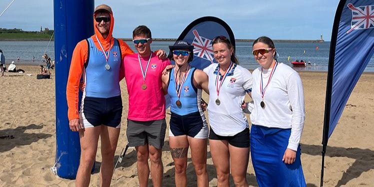 Five of the St Andrews rowing team stand smiling on a beach wearing medals and St Andrews team kit Five of the St Andrews rowing team stand smiling on a beach wearing medals and St Andrews team kit