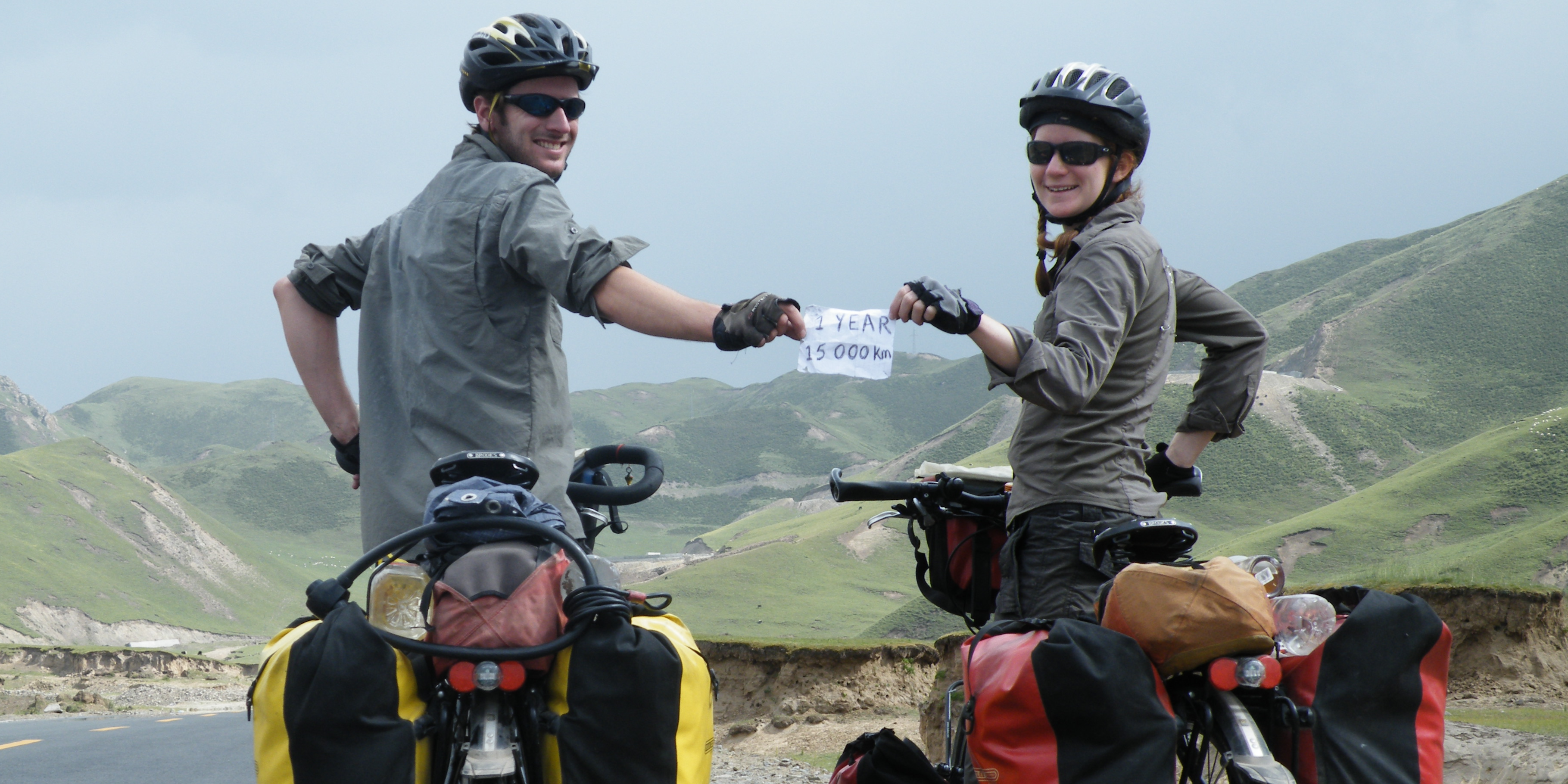Helen and Ed pose smiling for a photo on their bikes, holding a small paper sign that reads '1 year 15000 km'. The pair are wearing khaki shirts, bike helmets and dark sunglasses. A sprawling mountain range can be seen in the background of the image Helen and Ed pose smiling for a photo on their bikes, holding a small paper sign that reads '1 year 15000 km'. The pair are wearing khaki shirts, bike helmets and dark sunglasses. A sprawling mountain range can be seen in the background of the image