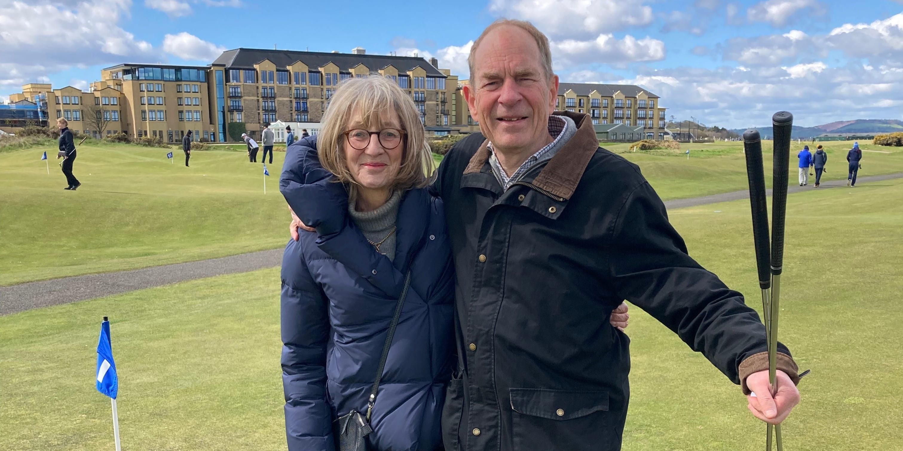 Anne and Patrick pose smiling for a photo on the Himilayas Course, St Andrews. The Old Course Hotel can be seen in the background of the image Anne and Patrick pose smiling for a photo on the Himilayas Course, St Andrews. The Old Course Hotel can be seen in the background of the image