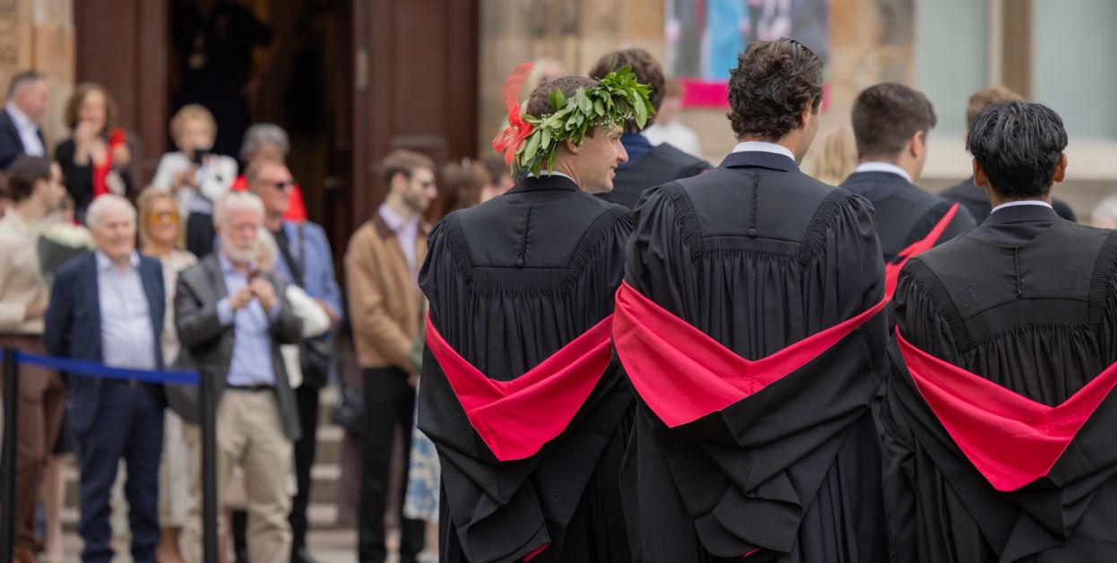 Two graduates hug during a graduation celebration. One graduate holds a bouquet of flowers wrapped in brown paper, while another bouquet is visible in the foreground.